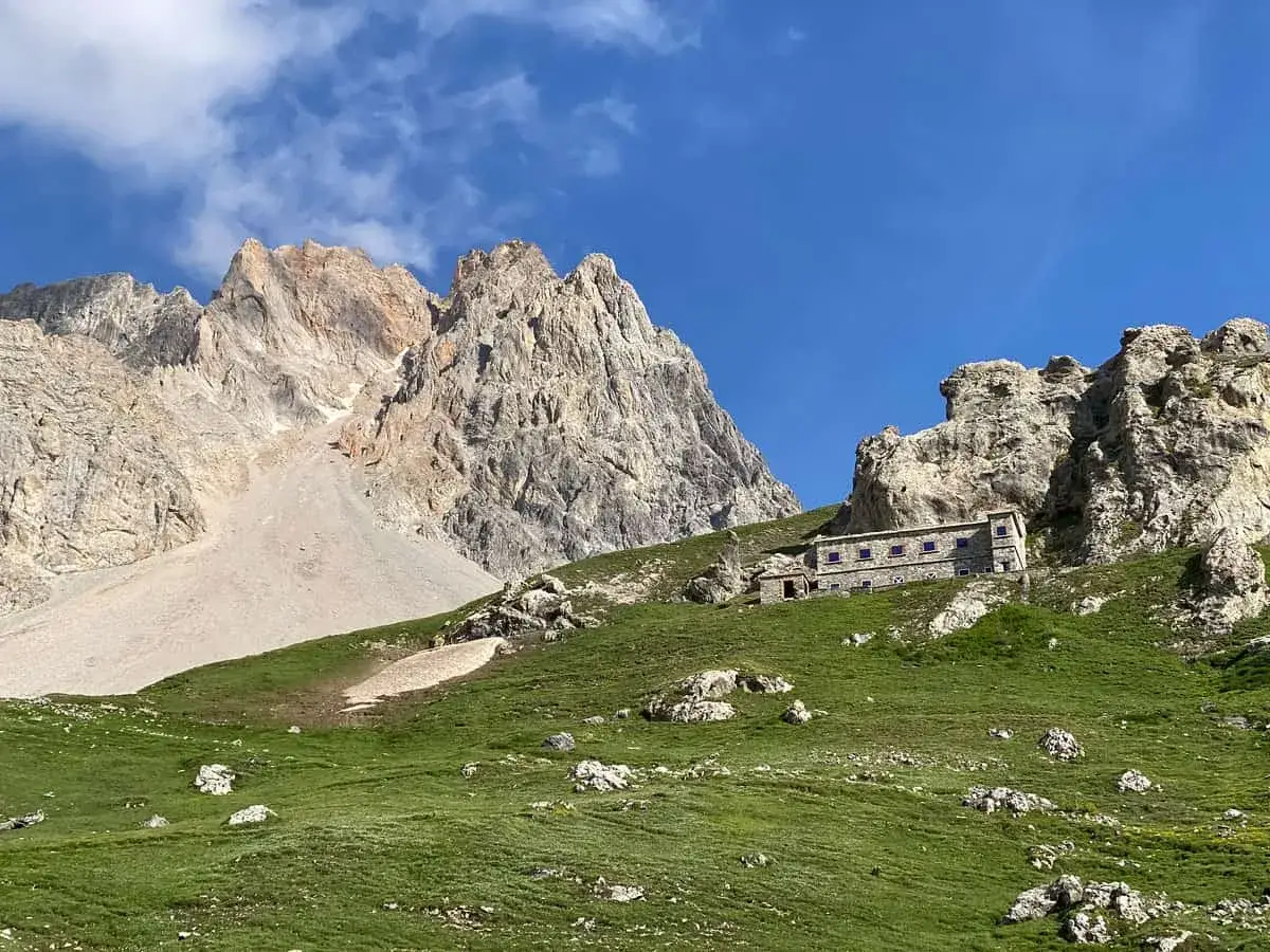 Rifugio alpino in pietra, incastonato nel verde di un prato in Val di Susa, dove si trova, con imponenti montagne rocciose sullo sfondo.