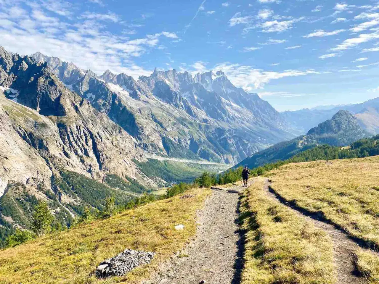 Sentiero panoramico tra i monti, ideale per itinerari in Valle d'Aosta. Un escursionista cammina su un prato dorato con vista su valli verdi e cime innevate.