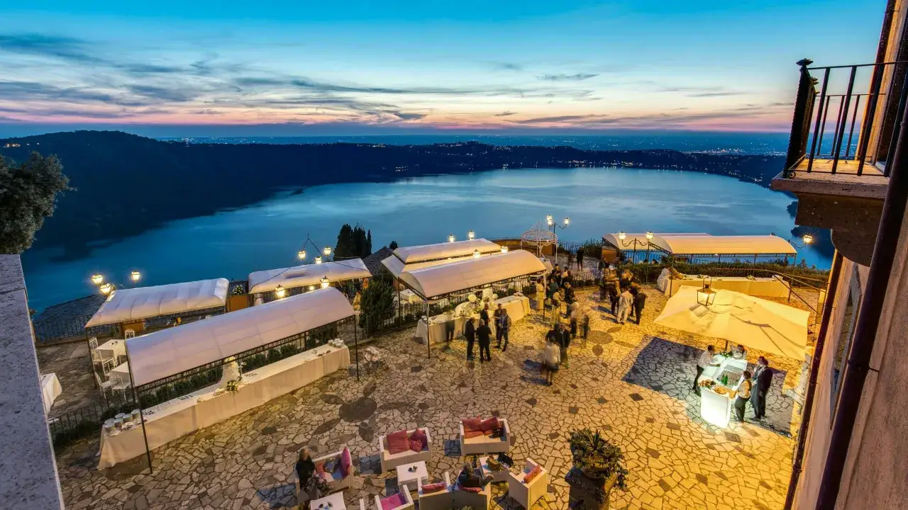 Serata elegante a Castel Gandolfo, Italia, con vista sul lago al tramonto. Tavoli allestiti e ospiti che socializzano.
