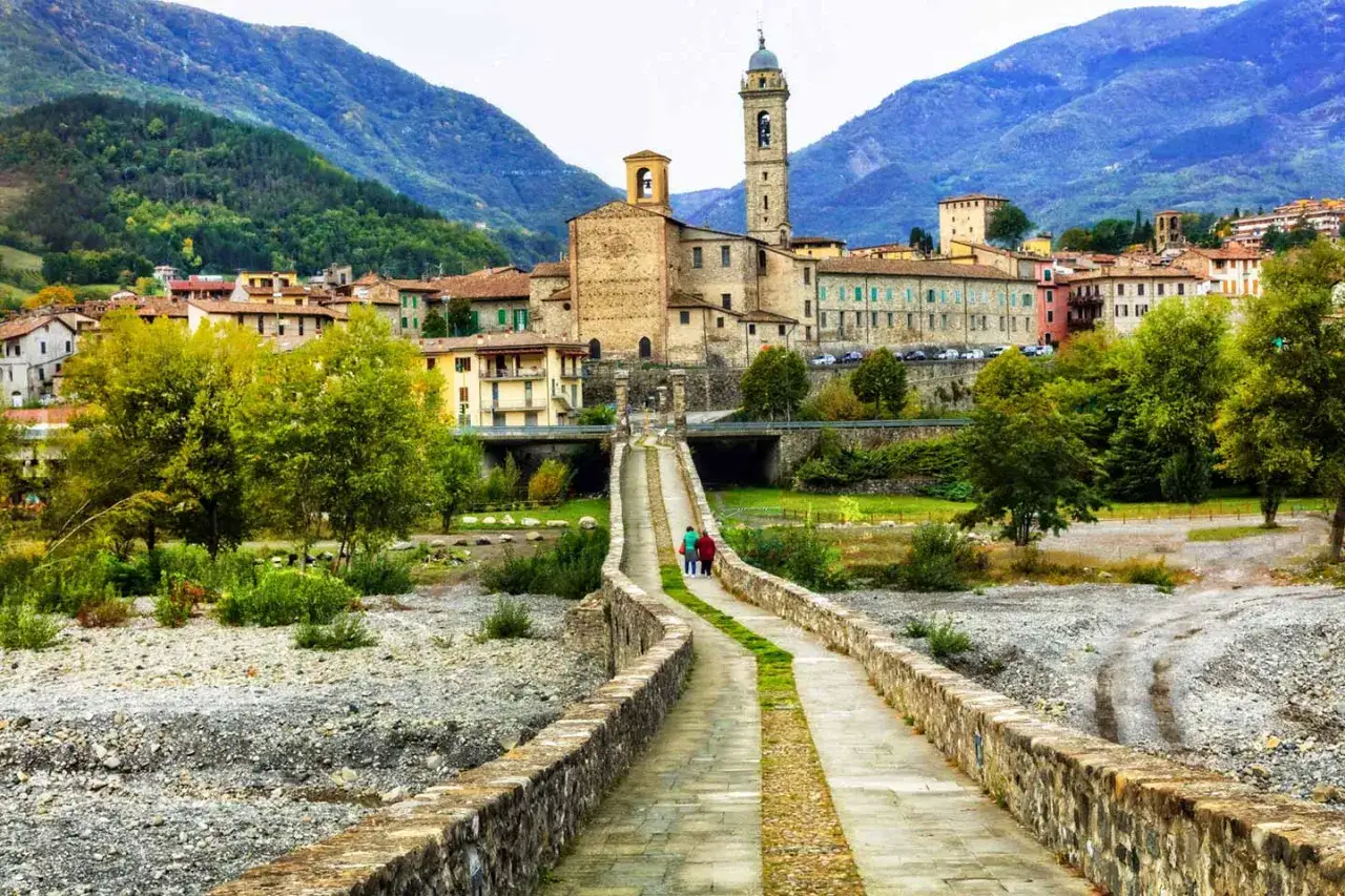 Ponte Gobbo e borghi vicino a Bobbio, con montagne sullo sfondo. Due persone passeggiano sul ponte antico.