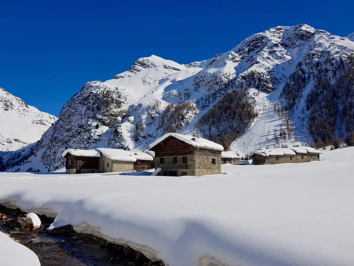 Case innevate nel Val di Rezzalo, con casolari in pietra e legno sotto montagne imponenti e un cielo azzurro intenso.