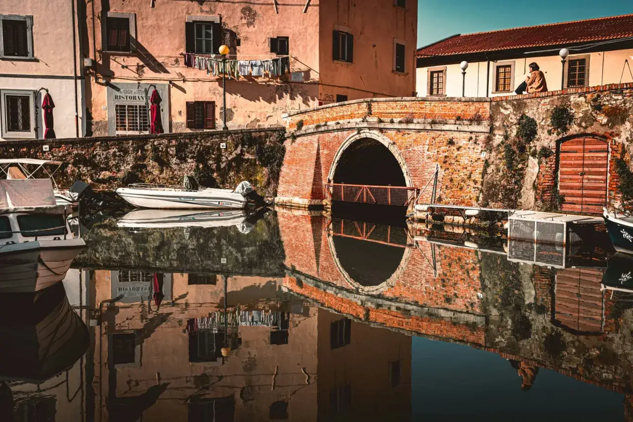 Un ponte in mattoni rossi riflette sull'acqua calma, con barche ormeggiate e case color pastello. Perfetto per gite di un giorno da Livorno.