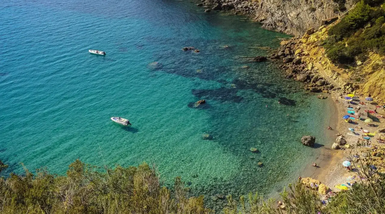 Spiaggia affollata con ombrelloni colorati e gente che prende il sole, bagnata da un mare cristallino toscana con barche ormeggiate.