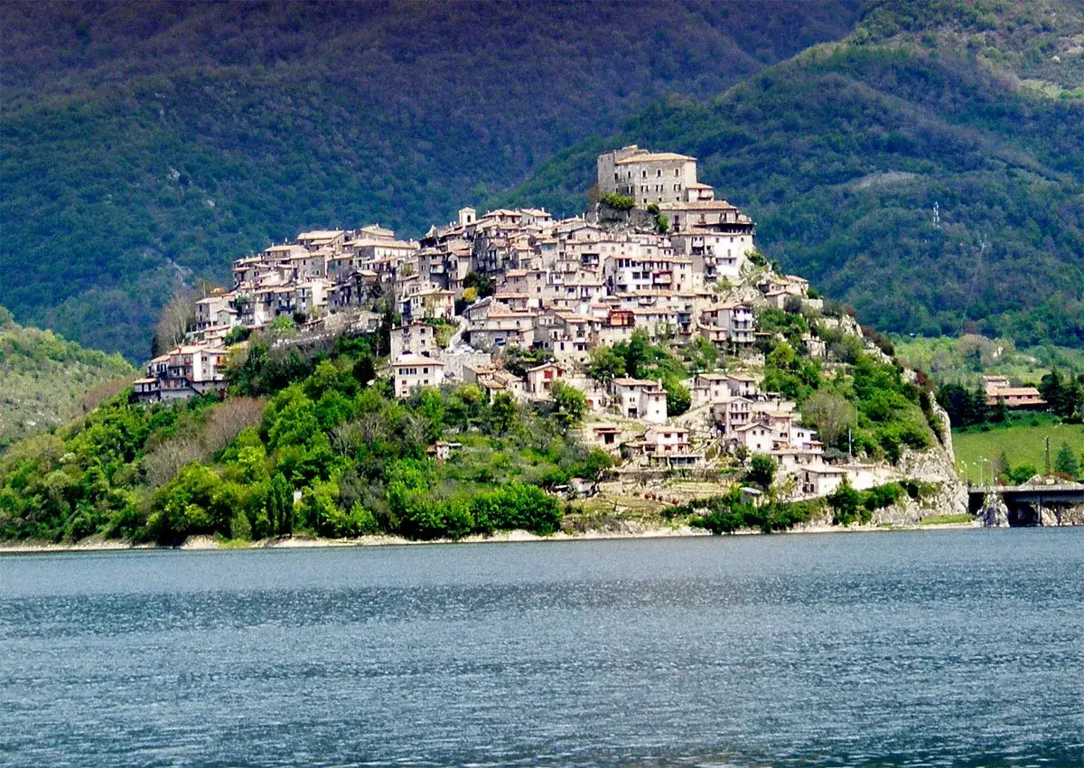Borgo medievale di Castel di Tora arroccato su una collina, con case in pietra e un castello in cima, affacciato su un lago azzurro.