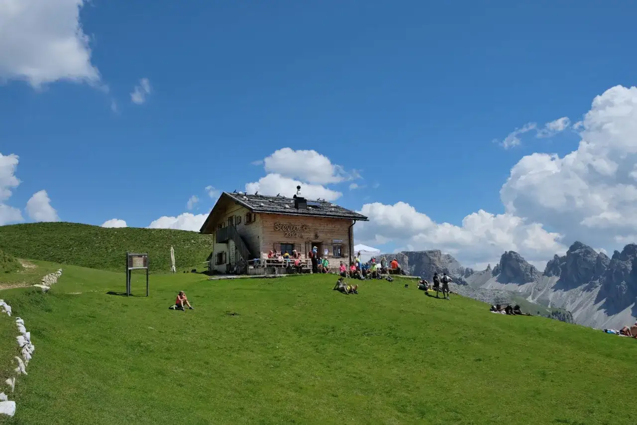 Rifugio Stevia, uno dei rifugi Val Gardena, accoglie escursionisti su un prato verde sotto un cielo azzurro con nuvole bianche.