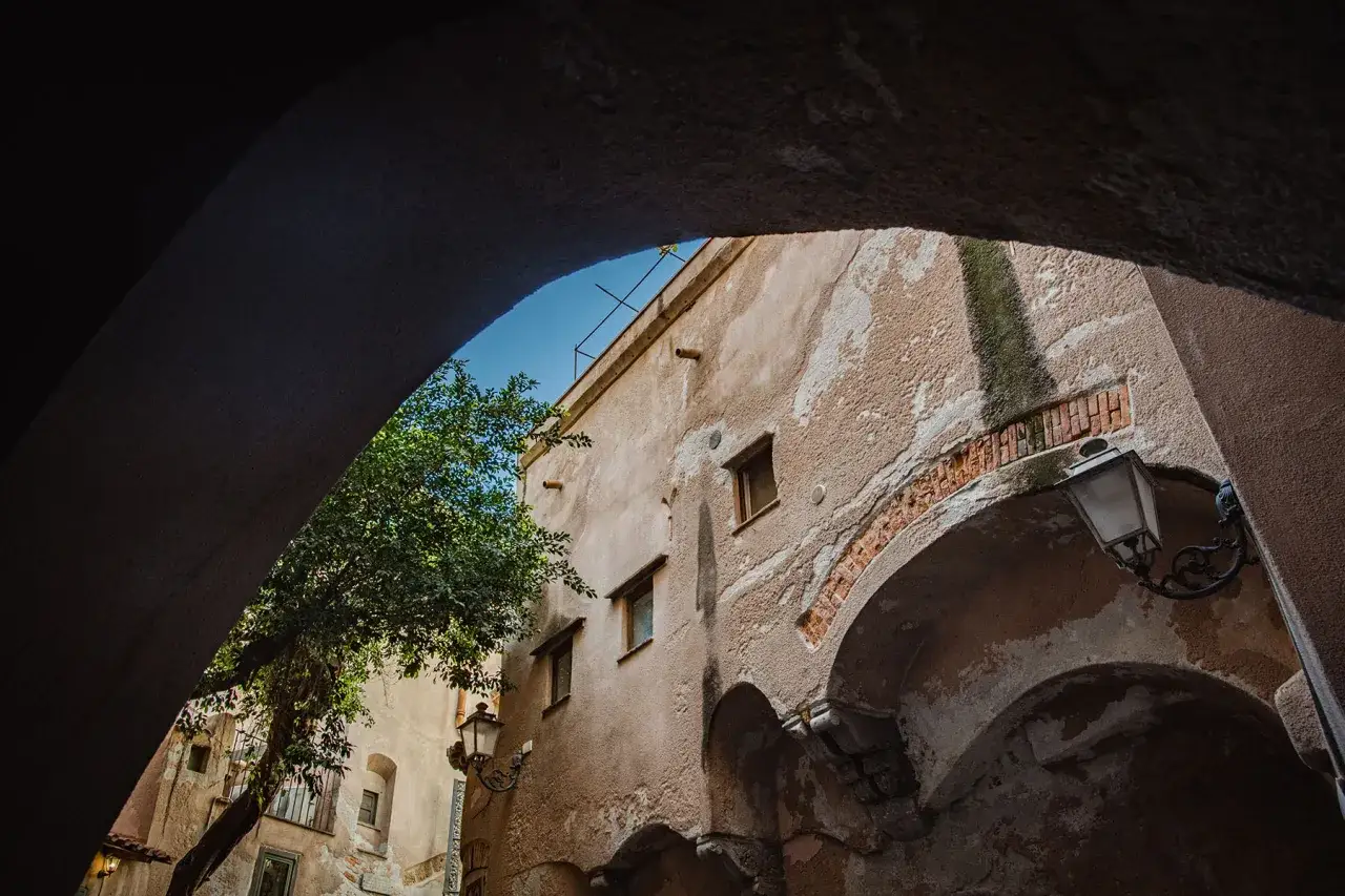 Un vicolo nel borgo Cefal&ugrave; centro storico, con un albero rigoglioso e un lampione antico sotto un arco.
