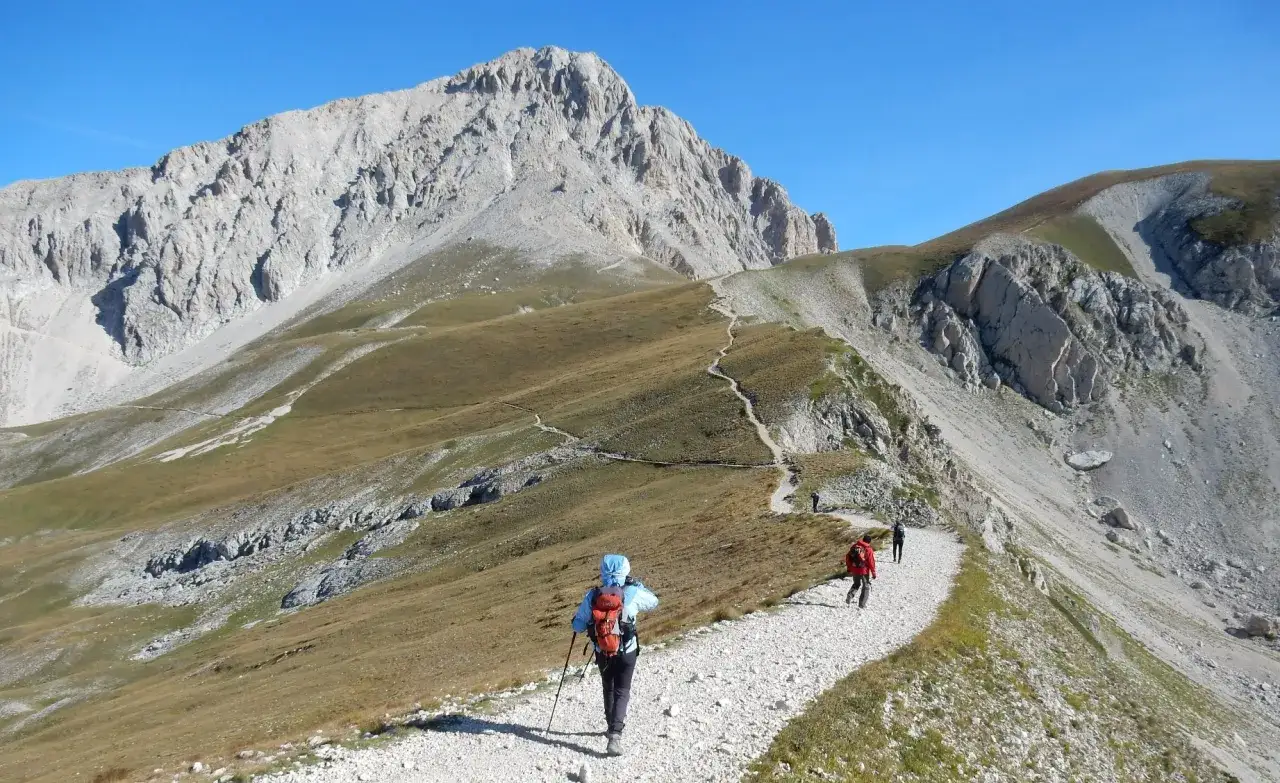 Escursione sul Gran Sasso: un sentiero serpeggia tra prati e rocce, con escursionisti che si godono il panorama mozzafiato.