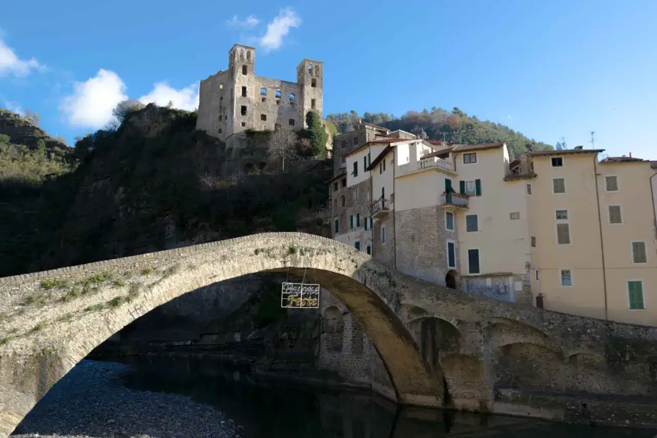 Antico ponte in pietra collega borghi entroterra ligure ponente. In alto, rovine di un castello dominano il paesaggio.