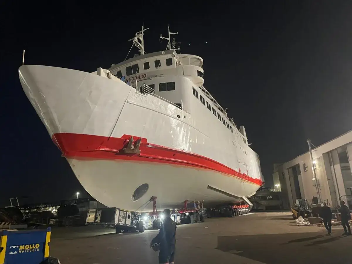 Il traghetto Isola del Giglio, bianco con fascia rossa, è sollevato su carrelli in un cantiere notturno.