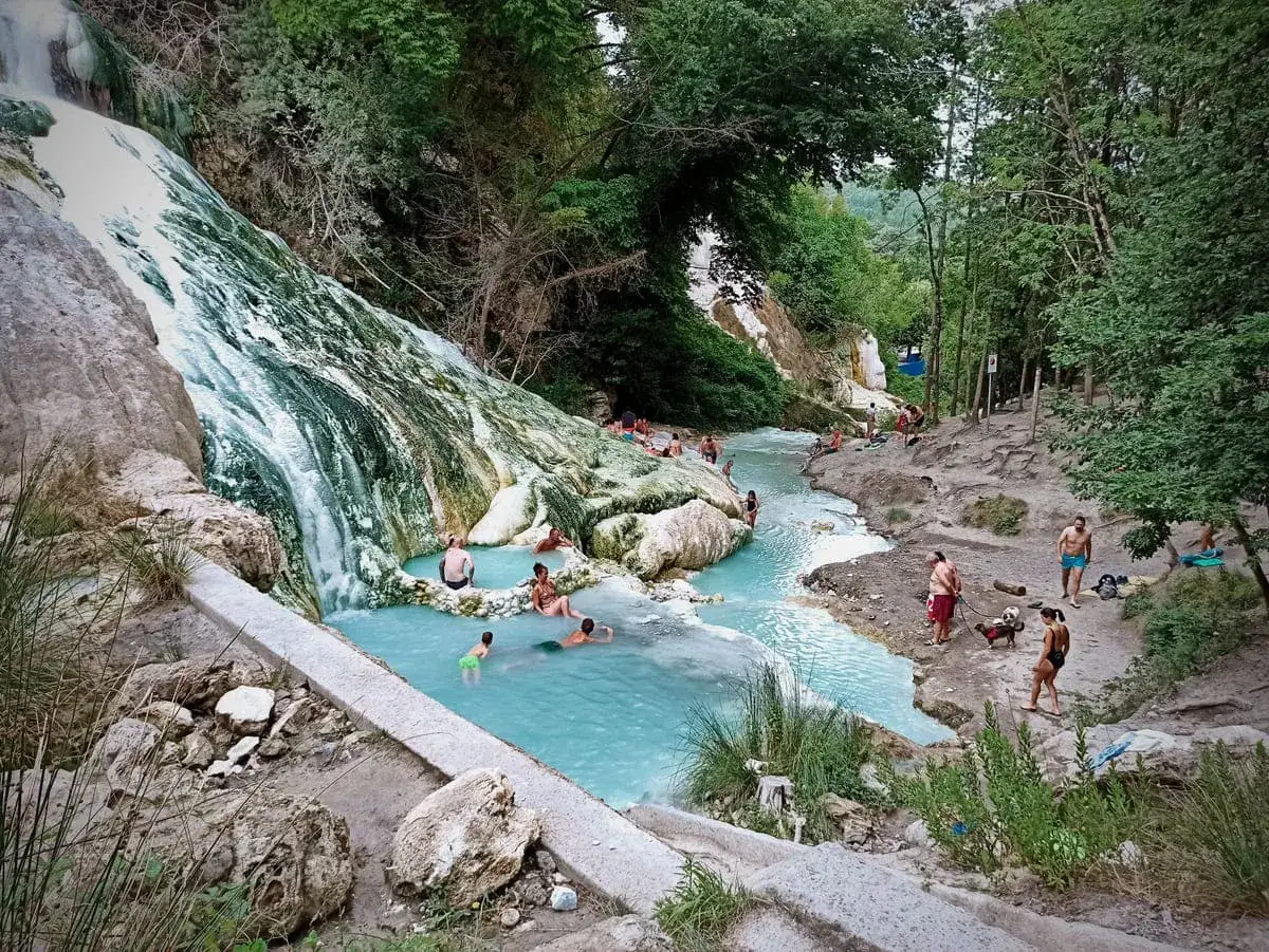 Cascata bianca e pozze turchesi ai Bagni di San Filippo, con persone che si rilassano nell'acqua termale.