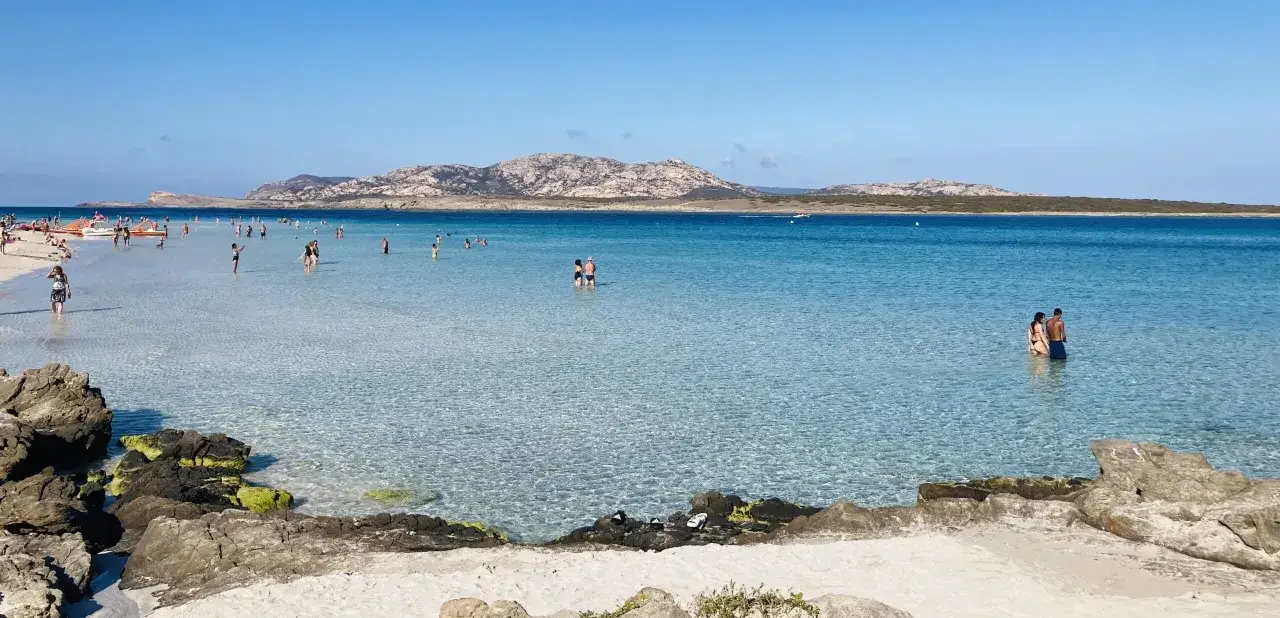 Acque cristalline e sabbia bianca: forse qual è la parte più bella della Sardegna? Gente che si gode il mare turchese, con montagne sullo sfondo.