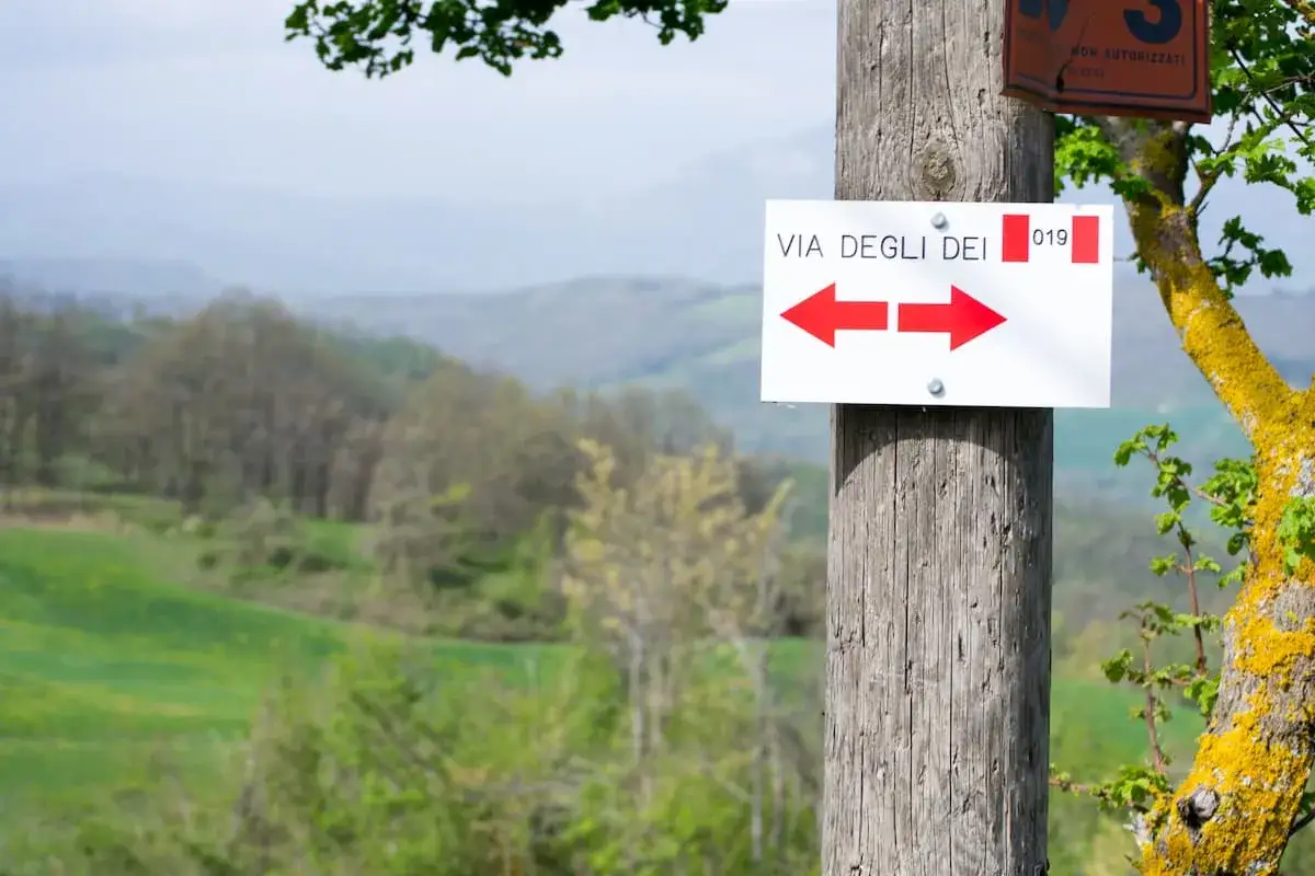 Cartello con frecce rosse indica le tappe della Via degli Dei. Paesaggio collinare sullo sfondo.