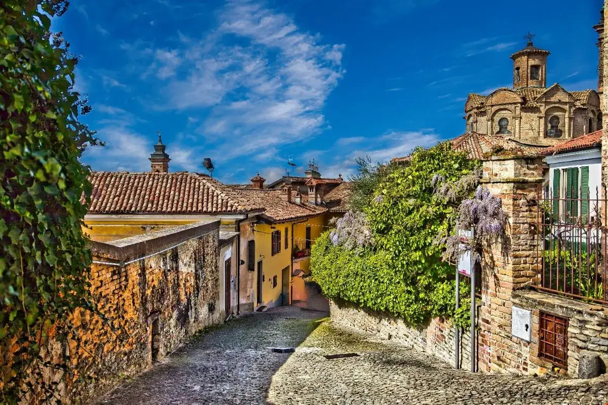 Un vicolo acciottolato nel centro storico di Neive, con case antiche, tetti in cotto e glicine in fiore sotto un cielo azzurro.