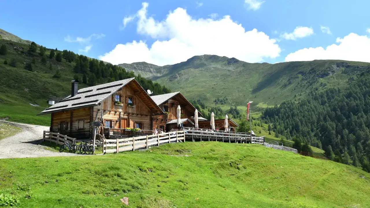Rifugio alpino in legno lungo il sentiero delle malghe, con tavoli all'aperto e montagne verdi sullo sfondo.