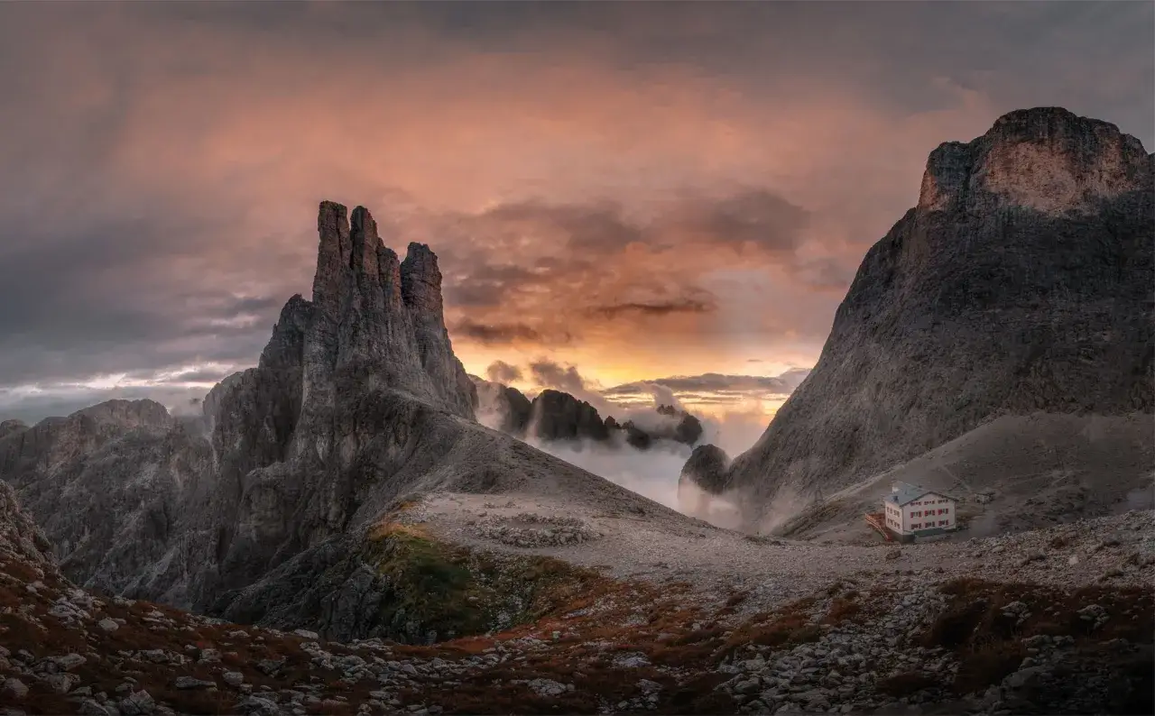Torri del Vajolet al tramonto, con nuvole infuocate e un rifugio solitario tra le rocce.