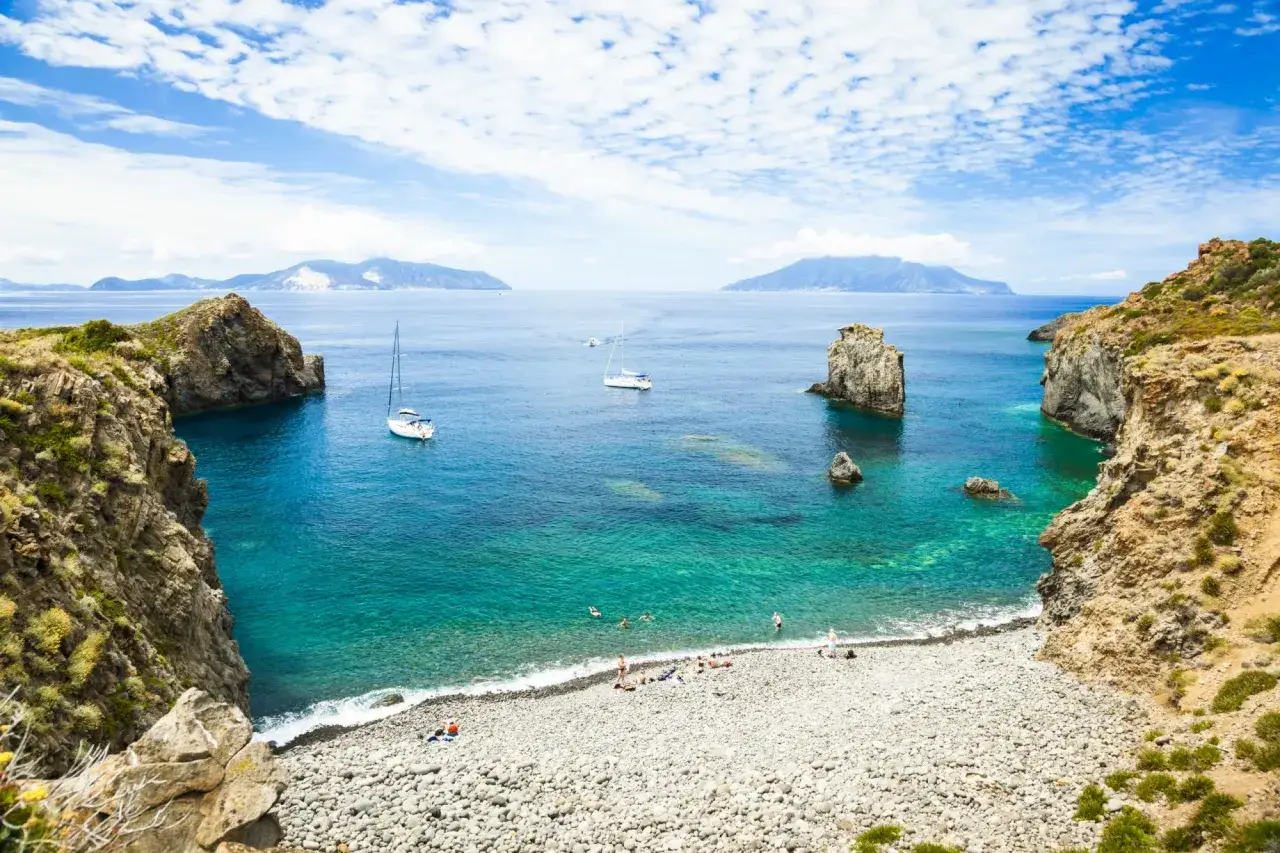 Spiaggia di ciottoli e mare turchese, ideale per scoprire come visitare le Isole Eolie in barca, con scogliere rocciose e isole all'orizzonte.