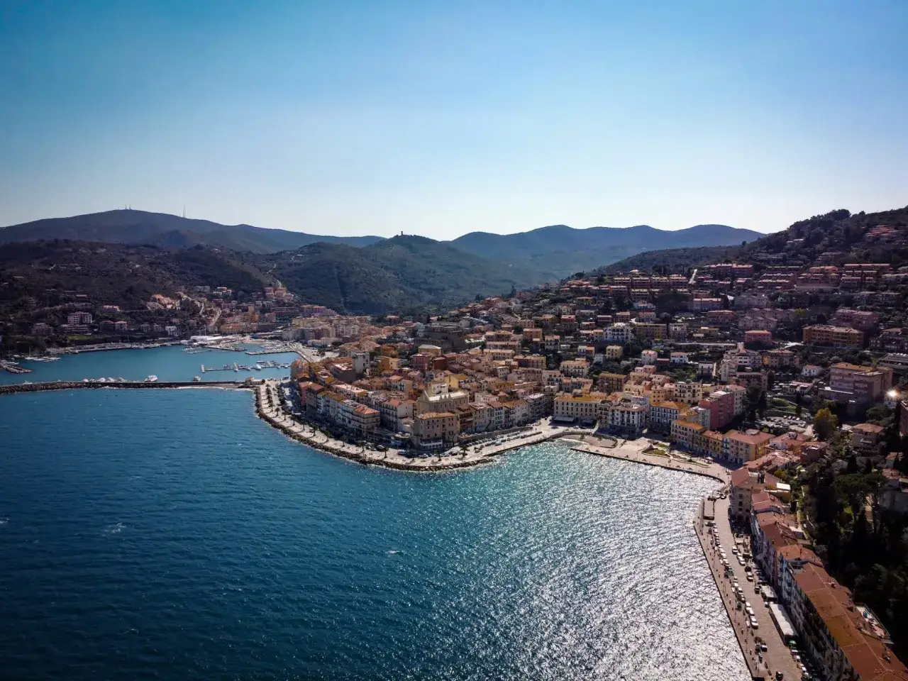 Vista aerea di Porto Santo Stefano con il suo porto, le case colorate che si arrampicano sulla collina e le spiagge di sabbia che invitano al relax.