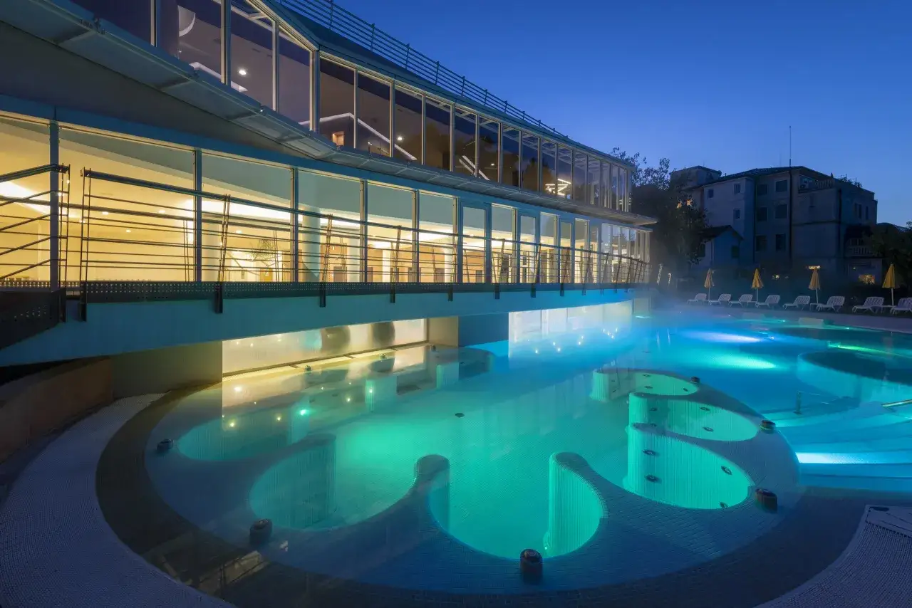 Piscina illuminata di verde e blu, con idromassaggi, sotto una struttura moderna. Un'oasi di relax che ricorda le antiche terme preistoriche a Padova.