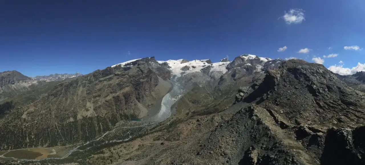 Vasto panorama alpino con cime innevate, un ghiacciaio che scende a valle e un torrente che serpeggia tra rocce e vegetazione. Il palon di Resy domina il paesaggio.