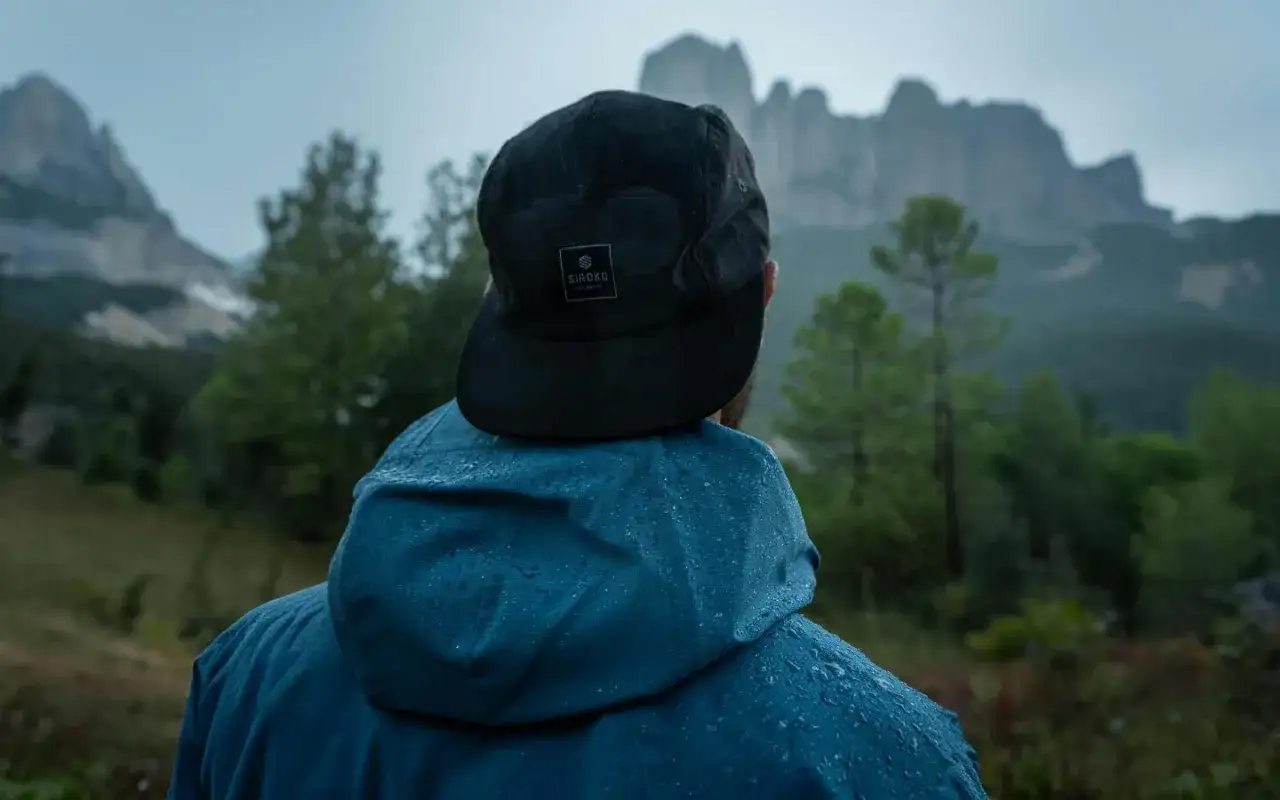 Uomo con cappellino nero e giacca blu impermeabile, pronto per l'abbigliamento montagna estate, con montagne sullo sfondo.