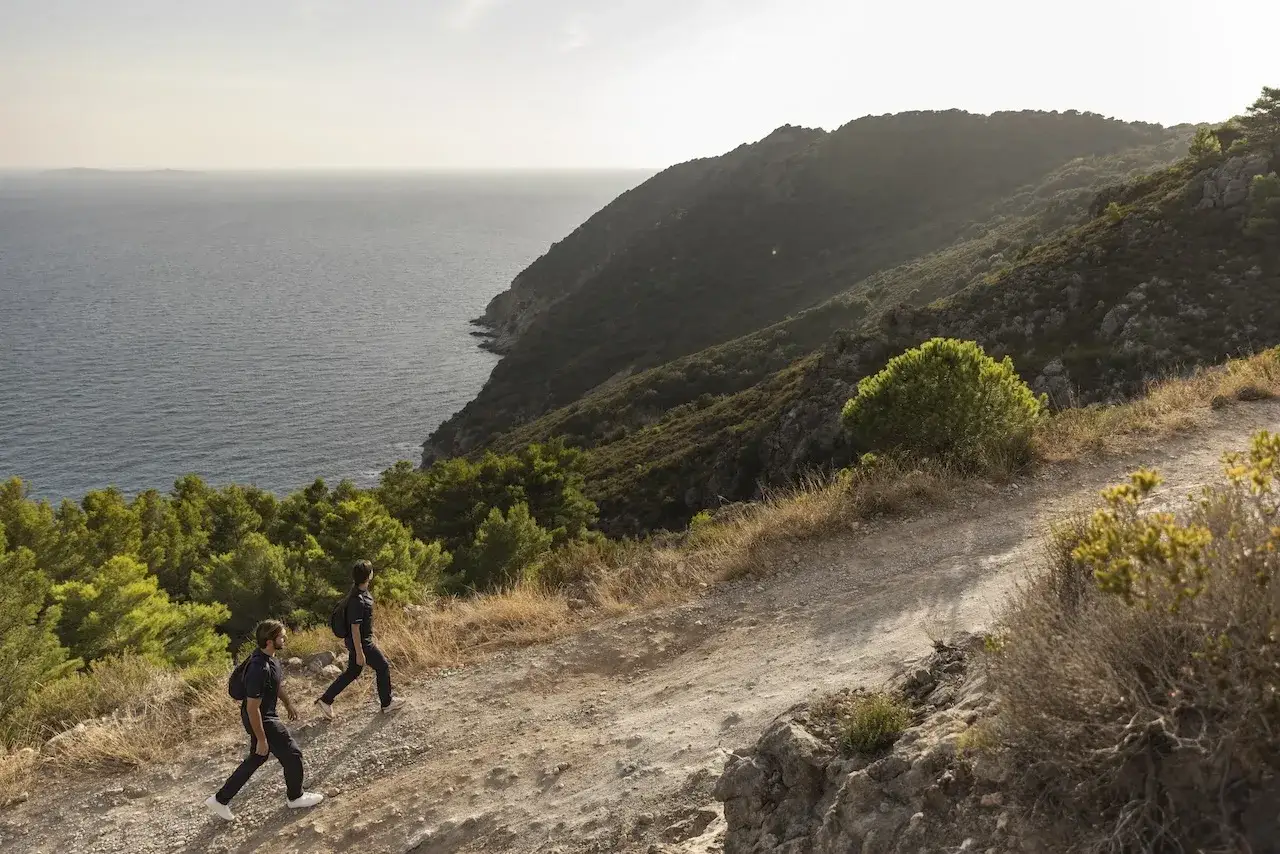 Due persone fanno cammini in Italia sul mare, su un sentiero panoramico tra macchia mediterranea e costa rocciosa.