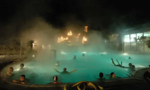 Persone si rilassano in una piscina termale notturna, con luci natalizie che brillano tra la nebbia, un'atmosfera magica nei bagni di Romagna.