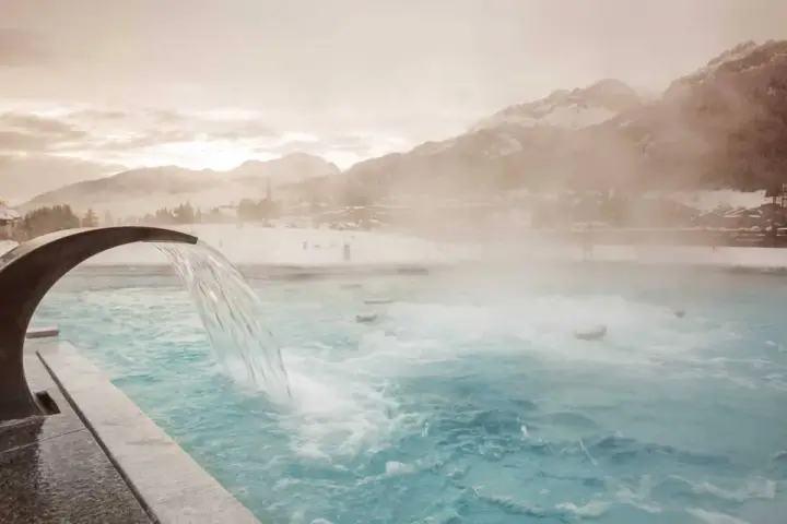 Una cascata d'acqua calda scende in una piscina all'aperto, con montagne innevate e un villaggio sullo sfondo. Un'oasi di relax a Pozza di Fassa QC Terme.