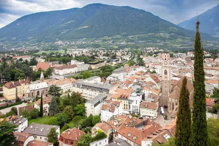 Vista aerea del centro storico di Merano, con montagne sullo sfondo e un campanile che svetta tra i tetti rossi.