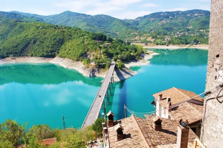 Panoramica mozzafiato del lago turchese vicino a Castel di Tora, con un ponte che attraversa le acque cristalline e montagne verdi sullo sfondo.