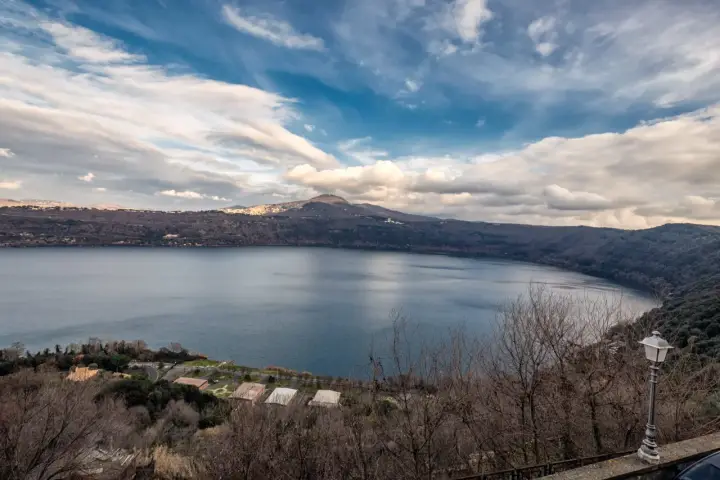 Vista panoramica del lago di Castel Gandolfo, Italia, con colline boscose e un cielo nuvoloso.