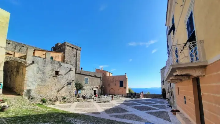 Piazza nel centro storico di Borgio Verezzi con edifici antichi, tavolini all'aperto e il mare sullo sfondo.